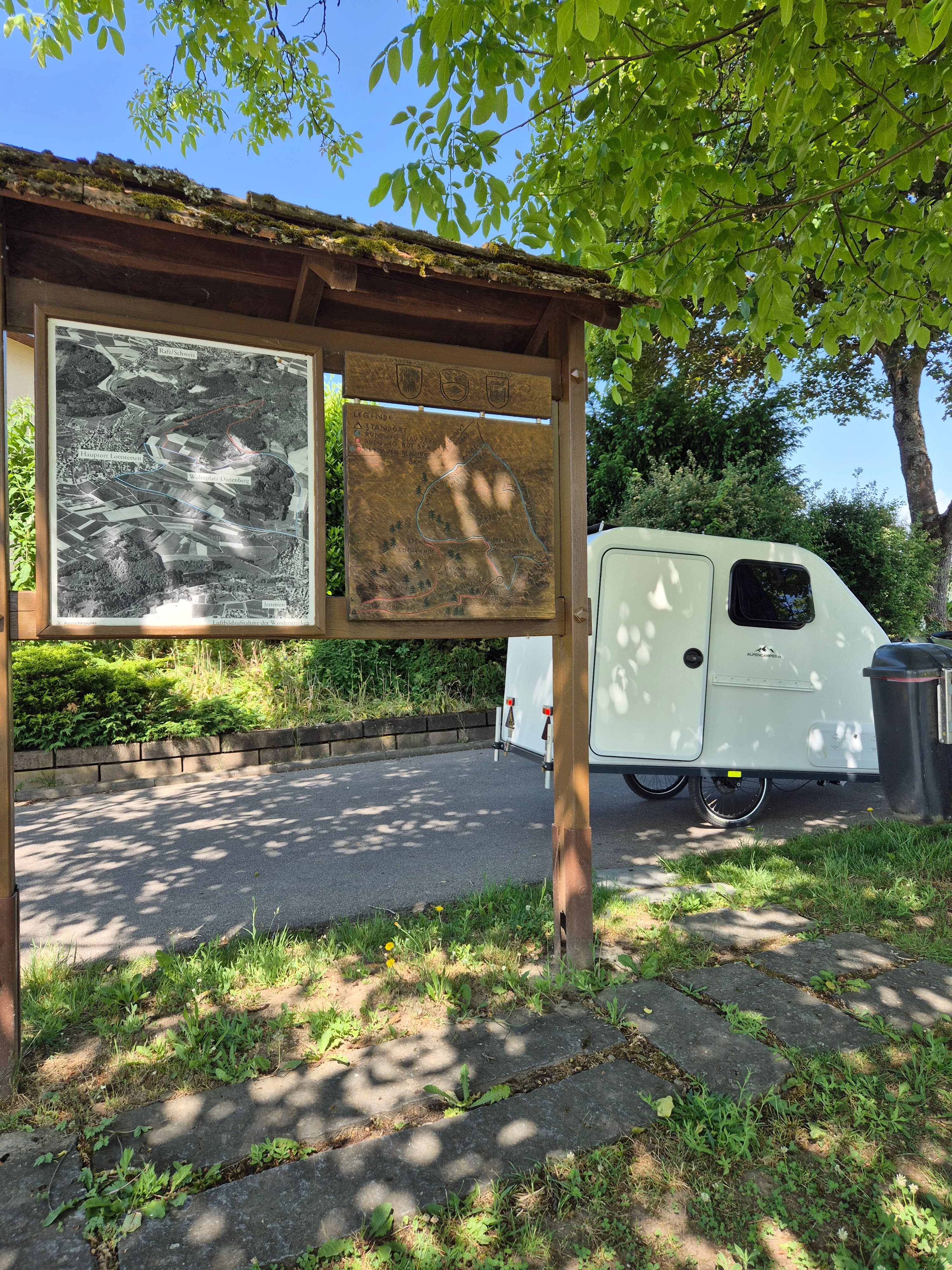 Der weiße Tiny-Camper steht am Wegesrand neben einer hölzernen Wanderkarte im Grünen